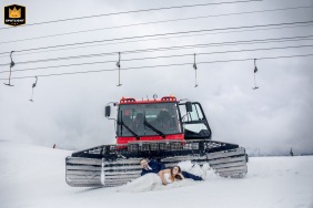 The bride and groom lie playfully in the snow in front of a snow groomer at Les Deux Alpes, France
